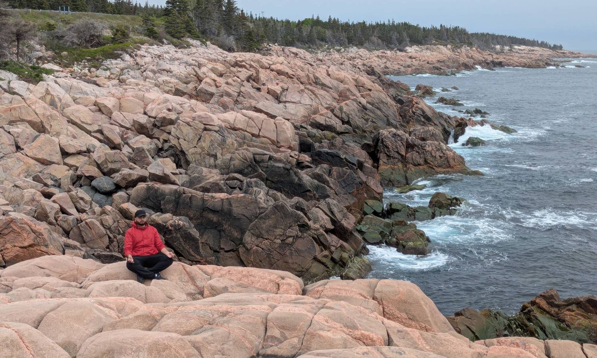 Shaun sitting cross-legged on pink granite rocks beside the ocean, with waves breaking along a rocky coastline.